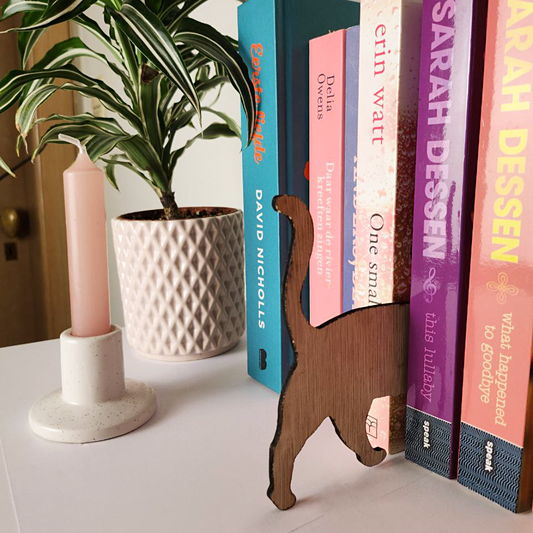 Een kattensilhouet dat uit hout werd gesneden staat tussen een rij boeken in. Het katje lijkt in de boeken te lopen, want je ziet enkel haar achterste poten en staart. 
