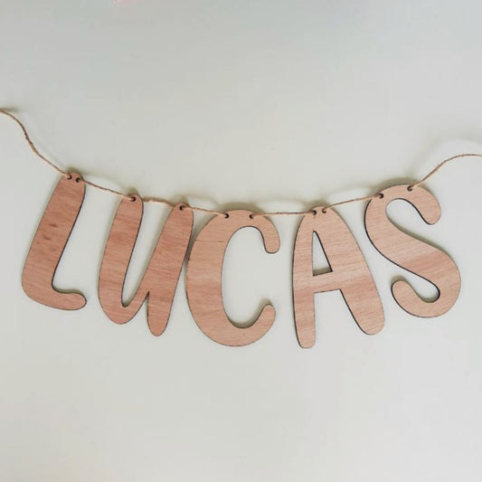 Houten slinger van uitgesneden letters die de naam 'Lucas' vormen. De letters zijn met elkaar verbonden door middel van een jute touw. 
